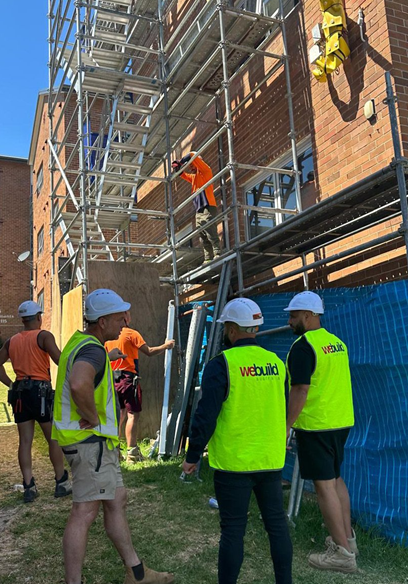 Bar Beach — We Build Australia site team in branded high-vis vests during a safety briefing beside scaffolded LAHC building